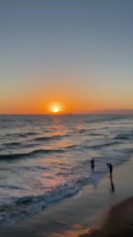 A wide shot of a beach during sunset. The sun is setting over the ocean, casting an orange glow across the sky and water. The waves are crashing on the shore, and two people are standing in the shallow water. One person is taking a photo of the sunset, and the other is looking out at the ocean. The beach is sandy, and there are some rocks in the distance. The sky is clear and blue, with a few clouds.