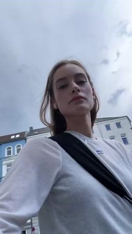 A woman stands outside on a cloudy day. She is looking down into the camera, then looks up to her right. She has fair skin, and is wearing a white long-sleeved shirt with a small blue label, a black cross-body bag, and a white wireless earbud in her left ear. Buildings are visible behind her.