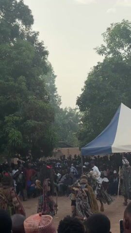 The video shows a vibrant outdoor gathering, likely a cultural event or celebration. A crowd of people of various ages is assembled in a clearing, with some seated under a blue and white tent and others standing around the perimeter. The event is framed by tall, leafy trees on either side, creating a natural enclosure. Several people in traditional attire with masks and costumes are dancing in the center of the crowd, their movements suggesting a performance or ritual. People are holding up phones to record the event. The general atmosphere is lively and celebratory.