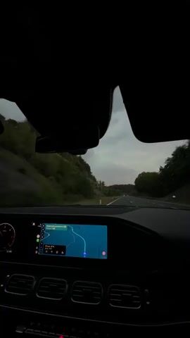 The video shows the interior of a car driving on a road with trees and hills on the side. The car's navigation screen is visible, showing a map with a blue route. The sky is cloudy and it appears to be dusk or early morning.