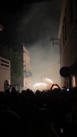 A crowd of people watch fireworks in a narrow street at night. The fireworks are bright and create a lot of smoke. Some people are holding up their phones to record the event. There are buildings on either side of the street, and some trees are visible in the background. The atmosphere is festive and exciting.