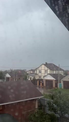 A low-angle, vertical shot captures a stormy sky above a residential area. Raindrops are visible on the camera lens, creating a blurry effect. In the foreground, a reddish-brown roof of a house is seen, with more raindrops on it. Beyond this, several other houses with pitched roofs are visible, many with attached garages. The sky is overcast with dark gray clouds, suggesting a heavy downpour. The street and yards appear wet, indicating recent or ongoing rain. Trees and fences are scattered throughout the scene. The overall impression is one of a stormy, wet day in a suburban or semi-rural setting.