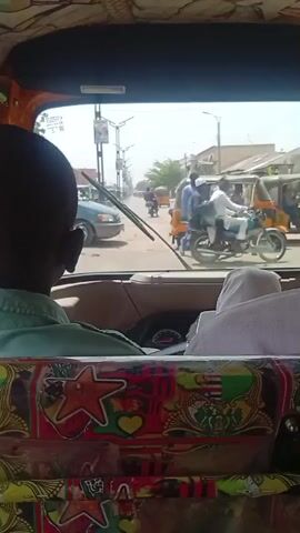 From inside a decorated auto rickshaw, the video shows a sunny street scene in an urban area. Several auto rickshaws, motorcycles, and bicycles are visible on the road along with pedestrians. The rickshaw is moving, and the perspective is from the rear. A passenger in the rickshaw is visible in front, and the interior is decorated with colorful patterns.