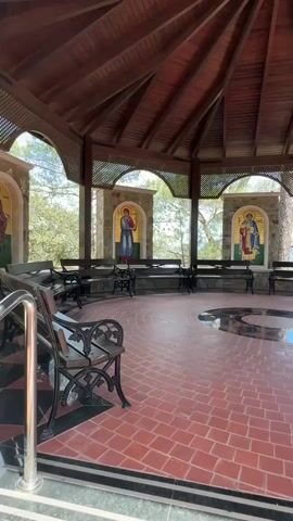 The video shows the inside of a circular gazebo-like structure. The structure has a wooden roof and arched openings revealing an outdoor landscape of trees. Along the arched openings are stone blocks and decorative latticework. There are religious mosaics set within these arches. Benches are arranged along the perimeter of the space facing inward. The floor is made of red clay tiles with a circular black and gray pattern in the center. A metal railing is visible in the lower left corner. The lighting is bright and natural.
