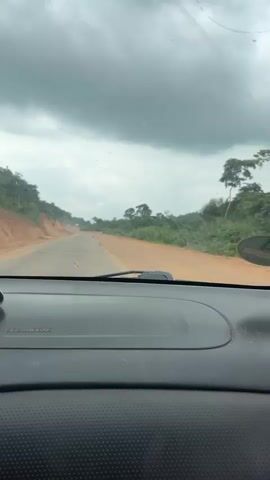 A first-person perspective video shows a road trip through a rural landscape. The view is from the inside of a car, looking out the windshield. The road ahead is paved but appears to be in poor condition, with visible cracks and uneven surfaces. The surrounding environment is lush and green, with dense vegetation and trees on either side of the road. The sky is overcast with heavy, dark clouds, suggesting the possibility of rain. The dashboard of the car is visible in the foreground. The video captures the feeling of traveling on a remote or underdeveloped road.