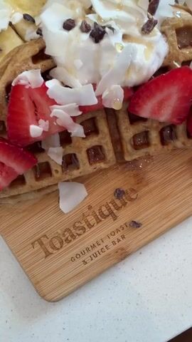The video opens with a close-up shot of a waffle topped with whipped cream, sliced strawberries, banana slices, coconut flakes, and chocolate chips. A golden fork and knife are placed next to the waffle on a wooden board. The board has the logo "Toastique GOURMET TOAST & JUICE BAR" etched into it. To the left of the waffle, a glass bottle with a green label and a green liquid inside is visible. In the background, a person is seated at a table in a cafe-like setting, with other patrons and restaurant decor also discernible. The camera then pans slightly to reveal a green beverage in a clear plastic cup with a straw, and another glass bottle. The final scene shows a wider view of the cafe interior, with several tables and chairs, and patrons engaged in various activities.