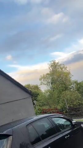 The video shows an outdoor scene with a cloudy sky. In the foreground, there is the side of a building with a dark gray wall and a brown roof. There are two vents on the wall. Further back, there is a wooden fence and trees. The camera angle is tilted, looking upwards.