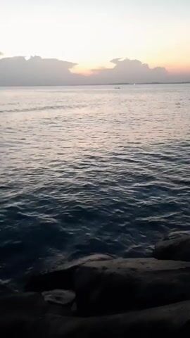 The video shows a calm body of water, possibly an ocean or a large lake, at dusk or dawn. The sky is pale with some clouds, and a hint of color suggests a sunrise or sunset. A rocky shoreline is visible in the foreground, and a small boat can be seen in the distance. The water has gentle ripples on its surface.