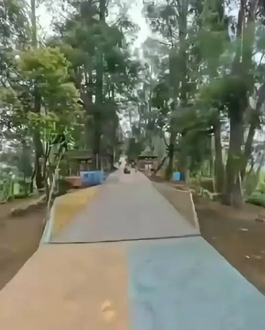 A first-person perspective video shows a paved pathway lined with trees. Small structures or stalls are visible on either side of the pathway. Several people are walking or standing in the distance along the path. The ground has areas of gravel and some construction materials. The overall environment appears to be an outdoor recreational area or park.