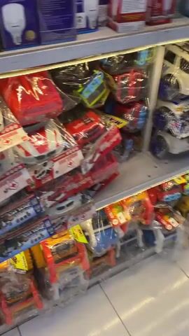 The video shows a display shelf stocked with various children's toys, predominantly toy vehicles. In the foreground, there are several rows of packaged toy cars and trucks, some appearing to be emergency vehicles like ambulances with red crosses. There are also toy police cars. Further back, a large number of toy train engines, resembling characters from children's shows, are densely packed on the shelf. The toys are all individually wrapped in clear plastic packaging, and some have colorful labels or tags attached. The background is slightly blurred but suggests a retail environment, possibly a toy store or a section of a larger store dedicated to toys. The lighting is bright, illuminating the products on display. There are no people visible in the video.