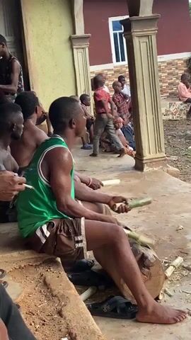 The video depicts an outdoor scene in Ogbu, Nigeria, featuring multiple individuals, primarily adult males and children.

The initial shot focuses on a man in a green tank top and brown shorts, seated on a wooden surface, rhythmically playing a large wooden drum with two sticks. He is smoking a cigarette. In the background, several children are gathered near a building with red and brown tiled walls and cream-colored pillars.

The scene then shifts to an open, unpaved dirt area surrounded by lush green foliage and trees. Several men are seen running and walking across this space. One man in a patterned shirt and dark pants runs past the camera. In the wider shot, other men are visible walking or standing, with one man crouching down. A group of people are gathered further in the background, standing and sitting under a large, leafy tree.

The ground is bare earth, suggesting a rural or village environment. The lighting indicates it is daytime, possibly overcast. No visible text is present in the video.