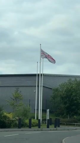 The video shows a Union Jack flag waving atop a white flagpole. Two additional flagpoles stand bare next to it. The flag is positioned in front of a large, industrial-looking building with a gray, corrugated metal exterior.  Some greenery including trees and shrubs are visible in front of the building. A road is visible in the foreground, demarcated by black posts. The sky is overcast.  The video appears to have been taken from inside a car, evidenced by the angle and slight movement. Loud music plays throughout the recording.