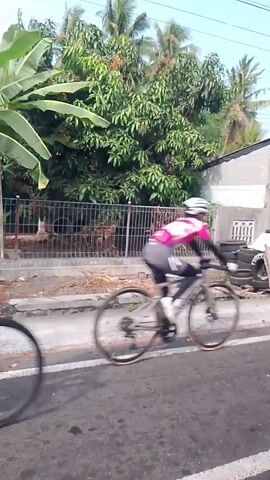 The video shows a street scene with several cyclists riding on a road. The cyclists are wearing helmets and cycling attire. The road is paved and has white lane markings. On the sides of the road, there are trees, houses, and a fence. In the background, there are power lines. The weather appears to be sunny. One cyclist in the foreground is wearing a red and white jersey, while another cyclist is wearing a green and white jersey. The cyclists are moving at a moderate pace. The video is shot from a moving vehicle, providing a dynamic perspective of the cycling event.