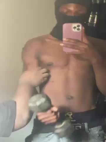 A shirtless man with a black ski mask stands in front of a mirror holding a dumbbell in his right hand and a purple smartphone in his left hand. The man's face is obscured by the ski mask, only his eyes are visible. He is holding his phone up to take a picture or video of himself in the mirror. In the background there is a light-colored wall and some items hanging on the wall.