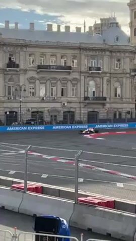 A Formula 1 car races on a track surrounded by barriers. The backdrop features a large building with arched windows and an American Express banner. The car is white with red and blue accents and "VISA" emblazoned on its side. The track is surrounded by spectators, and there is a large screen displaying race footage. The camera angle shifts slightly to show more of the track and cityscape, revealing trees and more buildings.