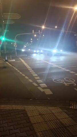 At night, a street intersection is depicted. Cars are driving through the intersection, their headlights illuminating the road and surrounding area. Traffic lights are visible, with one showing a green signal. Pavement markings indicate lanes and a bicycle lane. The scene is lit by streetlights, creating a bright and somewhat hazy atmosphere.