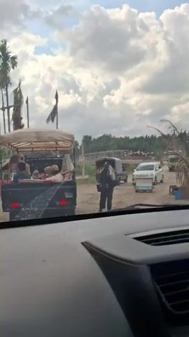 The video shows a view from inside a vehicle looking out at a dusty road. In the foreground, the dashboard of the vehicle is visible. Ahead, a small black pickup truck with a canopy is parked, carrying several people and what appear to be goods, possibly produce or supplies. To the right of the pickup truck, a person is walking away from the camera, carrying a bucket. Further down the road, there are other vehicles, including a white car and a green truck, and beyond them, trees and a bridge. The sky is partly cloudy. The scene appears to be in a rural or semi-rural setting, possibly a market or a roadside stop.