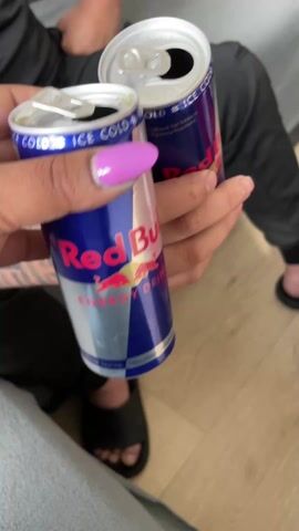 Two people are holding open cans of Red Bull energy drink. One person has purple nails. Both are wearing casual clothing and sandals. The scene appears to be indoors on a light wooden floor.