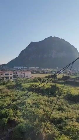 The video shows a clear blue sky over a rural landscape. In the foreground, there is green vegetation and some electrical wires. Several houses, which appear to be villas or vacation homes, are visible. In the background, a large mountain or hill dominates the horizon. The scene is peaceful and scenic, with a sense of a quiet, possibly coastal, environment. There are no people or animals visible in the video.