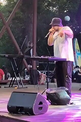 A man in a white t-shirt and a bucket hat stands on a stage, holding a microphone and gesturing with his hands. He is positioned behind two keyboards and other musical equipment. In the foreground, a large speaker rests on the stage. Behind the man, there is a wooden structure and trees, suggesting an outdoor setting. Other people are visible in the background, seated and standing, watching the performance. The stage is illuminated with purple lighting.