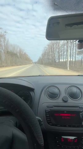 The video shows a driver's perspective of a car traveling on a straight, paved road. The road has a yellow center line and is bordered by trees on both sides. The trees appear to be bare, without leaves. The sky is cloudy. Inside the car, the steering wheel has a black, mesh-like cover. The dashboard is visible, with air vents and a car radio displaying 87.5 MHz. A rearview mirror and an air freshener hang from the windshield. Another car is visible in the distance. The car appears to be moving forward.