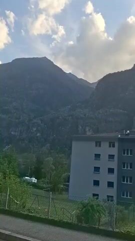 The video begins with a view of a mountain range under a cloudy sky. In the foreground, a building with a gray and white facade stands. The camera pans to the right, revealing more of the mountain landscape and a small town with buildings and a road. The sun is visible behind the mountains, casting a warm glow. The video appears to be shot from a moving vehicle, as indicated by the slight blur and the changing perspective. The environment is mountainous and rural, with signs of human habitation in the form of buildings and infrastructure. The time of day appears to be late afternoon or early evening, given the quality of light. There are no visible people or specific actions occurring in the scene.