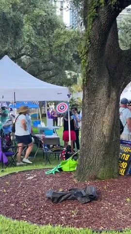 This media file captures an outdoor gathering in Fort Lauderdale, USA, likely during the daytime given the bright lighting. The scene is set in a park-like area with a large, mature tree prominently featured in the foreground. Beyond the tree, several white tents are visible, suggesting a planned event such as a rally, festival, or market.

Numerous people are present, engaged in various activities. Some are standing in small groups, conversing, while others appear to be observing or participating in the event. In the middle ground, there's a person sitting in a blue wagon, looking somewhat relaxed or perhaps tired. Closer to the camera, a group of people are gathered around signs, possibly engaging in a discussion or protest. One sign is partially visible with the text "TRUMP BUS GRASSROOTS" and another sign reads "MARCH FOR PEOPLE" with a logo that appears to be " MARCH FOR PEOPLES." Another sign in blue has text including "UNITED WE STAND TOGETHER WE MARCH." There are also American flags visible, indicating a potentially patriotic or political gathering.

A large screen or stage area is visible in the background, with more people gathered around it, suggesting a focal point for the event. The setting includes well-maintained green grass and scattered trees, with buildings in the distant background hinting at an urban environment. The overall atmosphere appears to be active and engaged, with a sense of community or shared purpose among the attendees. The weather seems fair, with no indications of rain or extreme conditions.