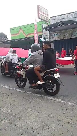 A procession is moving down a street. Two people are riding on a motorcycle, the driver wearing a helmet and a grey shirt, and the passenger wearing a black hoodie. In the background, a group of people are carrying a large red and white flag. Some of them are wearing green outfits and headscarves. They are walking alongside the street. Other people are visible in the background, and there are buildings and signs along the road. Another motorcycle is visible on the left side of the frame, also with two people riding it. A yellow object with writing on it is partially visible in the foreground on the left. The ground is covered in gravel.