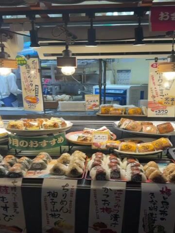 The video presents a static, eye-level shot of a food stall counter, showcasing an array of prepared Japanese food items. In the foreground, multiple rows of various onigiri (rice balls) and other small snacks are meticulously arranged on tiered, dark trays. Each section of food is accompanied by small white placards displaying Japanese text and prices, such as ¥280 and ¥210. Above the food displays, several larger signs with Japanese writing are mounted, one of which includes English text stating, "The next restroom is on the left." The display area is brightly lit by multiple industrial-style pendant lights suspended from the ceiling. In the background, behind a glass partition, a kitchen or preparation area is visible, where a person wearing a white uniform is partially seen standing on the left side of the frame.