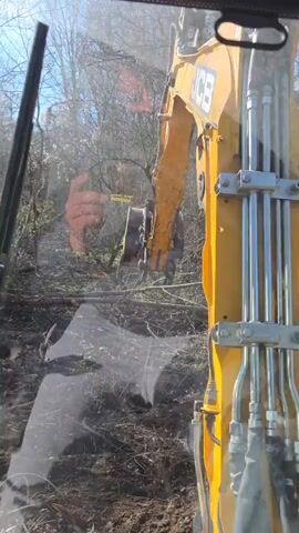 The video shows a dug trench in a field from the perspective of inside a piece of heavy equipment.  The field has dry vegetation and dirt. There are trees in the background and a bright blue sky with some clouds visible at the top of the frame. The view transitions from behind a dirty windshield to an unobstructed view, then back inside the cab of the heavy equipment.