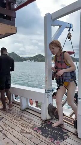 Several people prepare to dive off a boat on a cloudy day. One woman with yellow hair, wearing a yellow bathing suit and life vest, walks along the side of the boat, holding a diving mask. She stops next to another woman wearing a white top, denim shorts, and a life vest. A third woman with a pink hat, sunglasses, a black top with long sleeves and shorts, looks down at her phone. Boats are visible on the water and an island is visible in the distance. A welcome mat is near the back of the boat.