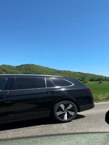 This video showcases a serene rural landscape under a clear, bright blue sky. In the foreground, a gray asphalt road with a white shoulder line runs horizontally, with a strip of green grass bordering it. Beyond the grass are vast fields, primarily a vibrant green, with some patches of lighter green and yellowish-green visible further in the midground, suggesting different types of crops.

Dominating the background are gently rolling hills, densely covered with lush green trees, creating a continuous forested expanse. The sunlight is strong, indicating a sunny day. The camera appears to be in a fixed position, capturing the landscape from a stationary or very slowly moving viewpoint, possibly from the side of a road or a slow-moving vehicle. The overall impression is one of peaceful natural beauty and wide-open spaces. No people or specific identifying objects are visible.