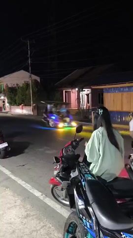 The video shows a nighttime scene on a street with several motorcycles. In the foreground, a person wearing a light green hoodie and a helmet is seated on a red motorcycle. Another motorcycle, partially visible, is in front of it. Further down the street, a white truck is parked, and several other motorcycles are visible, some with riders. Buildings line the street, illuminated by streetlights and lights from the buildings themselves. The atmosphere appears to be casual, possibly a gathering spot or a roadside scene.