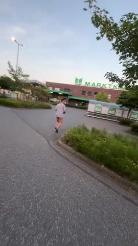 The video shows a person running in front of a "Marktkauf" supermarket. The person is wearing a grey t-shirt and plaid shorts. They are running away from the camera towards the entrance of the supermarket. In the background, there are several shopping carts neatly arranged under a shelter. The supermarket building is made of red brick with green trim and large green lettering spelling out "MARK TKAUF". There are also windows and a parking sign visible. The scene appears to be set in the late afternoon or early evening, with soft lighting and a clear sky, suggesting fair weather. The overall atmosphere seems ordinary, with a person engaged in a simple activity in a common public space. The text "MARK TKAUF" is prominently displayed on the building.