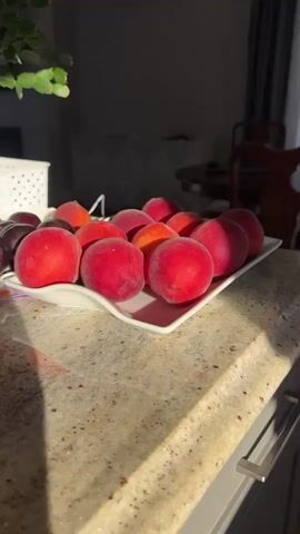 A close-up shot shows a white, wavy-edged dish filled with ripe peaches. The dish is placed on a granite countertop. Sunlight streams in from the right, casting shadows on the fruit and the countertop. In the background, a shadowy room is visible, with hints of furniture.