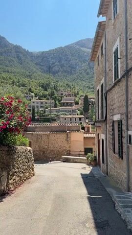 A narrow, paved road winds through a sun-drenched village nestled at the base of a lush, green mountain. On the left, a rough stone wall is adorned with vibrant pink flowers, their blossoms spilling over the top. Beyond the wall, more stone buildings are visible, some with terracotta roofs. The road curves towards the center of the frame, leading to a cluster of buildings. In the distance, houses and buildings ascend the mountainside, interspersed with trees. A tall, slender antenna or tower stands prominently on the slope. On the right side of the road, a stone building with dark shutters on its windows rises, casting a long shadow onto the pavement. The sky is a clear, bright blue, indicating a sunny day. The overall scene depicts a charming, rustic European village in a mountainous landscape.