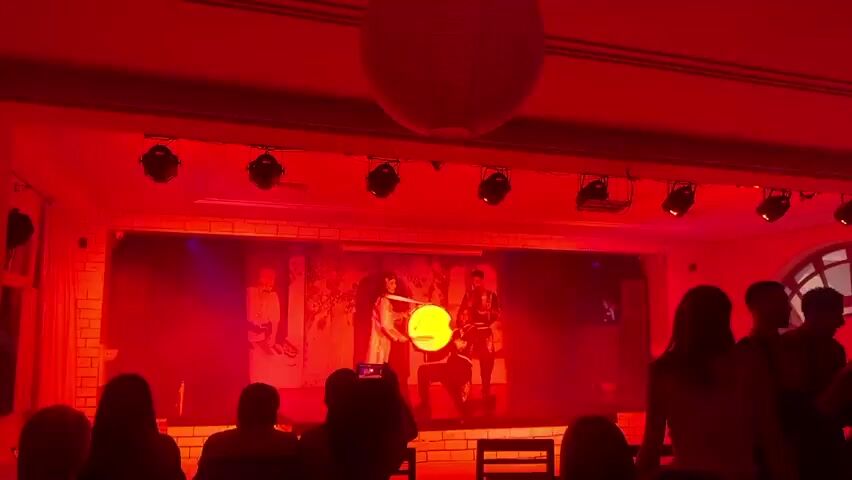 The video shows a performance taking place on a stage in a room lit with intense red lighting. The stage has a backdrop and is illuminated by spotlights. A large, round paper lantern hangs from the ceiling above the audience. On stage, at least two figures are visible, one appearing to play a large, illuminated drum-like instrument. In front of the stage, a crowd is watching the performance. Several individuals are holding up phones, seemingly recording or taking pictures. The environment appears to be a venue like a banquet hall or a performance space, characterized by architecture with features such as brickwork.