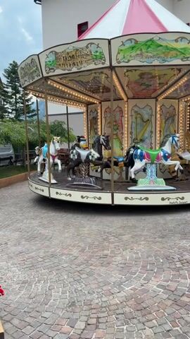 A small carousel is rotating outdoors. The carousel is decorated with various paintings, including landscapes and figures. There are several carousel horses, some of which are occupied by people. The ground around the carousel is paved with cobblestones. The carousel is illuminated by numerous small lights. In the background, there is a building and some greenery.