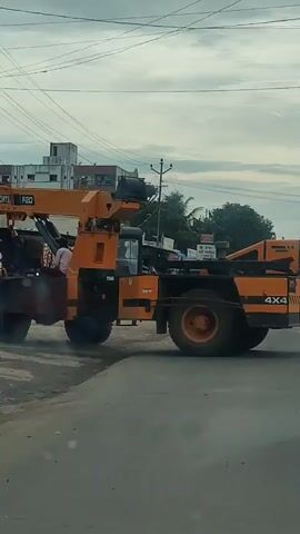 The video shows a yellow crane truck driving down a street. The truck has the marking 4x4 on the back. There are buildings and shops on the side of the road. A few people are standing near the shops. The sky is cloudy.