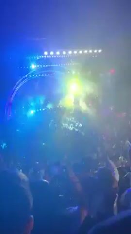 Here's a description of the video:

The video shows a concert with a large crowd of people. There is a stage with performers on it, visible at times through bright lights and colorful effects. Some of the shots are shaky due to being taken by someone in the audience. The lighting changes throughout, ranging from blue and purple to white and yellow. People in the crowd are holding up their phones, presumably to record the event. The overall atmosphere is energetic.