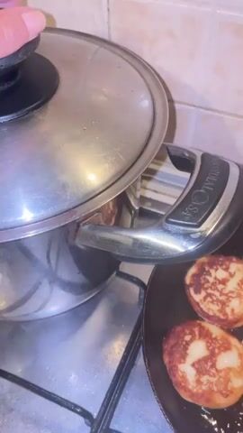 This close-up video shows a cooking scene in a kitchen. In the foreground, a black frying pan contains several golden-brown fritters, some with darker browned edges. The fritters appear to be sweet and fluffy. To the left, a silver pot with a black lid is simmering on a stovetop. Steam rises from the pot, suggesting it contains a hot liquid or food. The lid is slightly ajar, revealing a dark red mixture within, possibly fruit or vegetables being cooked. The stovetop is a metal gas hob with visible grates. The background shows a light-colored tiled wall, common in kitchens. The lighting suggests it is daytime. The overall atmosphere is domestic and focused on food preparation. The visible text on the pot handle reads "IONAL GASA," likely part of a brand name. The action suggests the preparation of a meal, possibly involving both sweet and savory components given the presence of fritters and the simmering pot. The scene is set in a kitchen in Castelfranco di Sotto, Italy.