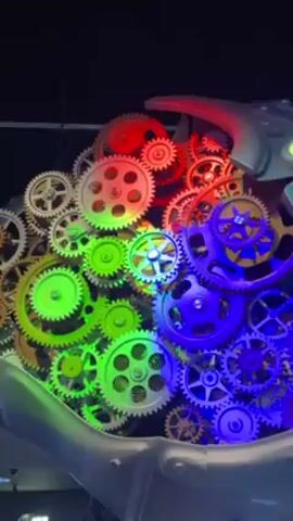 The video shows an elaborate mechanical display featuring numerous gears arranged in a dense cluster. The gears are illuminated with a spectrum of vibrant colors including red, green, blue, and white, creating a visually striking effect. The structure that houses the gears appears to be made of a silver material, resembling part of a machine or robot head. It has a smooth, metallic finish. The background is dark, highlighting the bright colors of the gears. The overall impression is of a complex, colorful mechanical system.