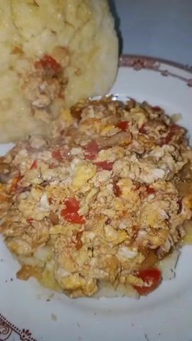 The video shows a plate of scrambled eggs with diced tomatoes and onions, served alongside a round, flatbread that appears to be a arepa. The arepa has a buttery sheen, suggesting it may be greased. A hand is seen lifting the arepa to create a sandwich with the scrambled eggs. The plate is white with a brown, patterned border. The surface underneath the plate appears to be a table, possibly white or light-colored.