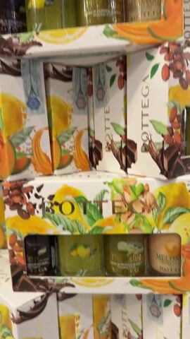 The video shows a close-up, slightly shaky, view of several boxes of Bottega products stacked on top of each other. The boxes are decorated with a colorful watercolor-style illustration featuring lemons, cantaloupe, pistachios, and chocolate. Each box has a rectangular window through which small jars of what appear to be creams or lotions can be seen. The labels on the jars are in Italian. The lighting suggests an indoor setting, possibly a store or warehouse. There are no people visible in the video.
