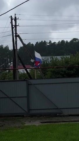 A Russian flag is seen attached to a flagpole. The flag is positioned behind a grey metal fence. Above the flag and fence, there are electrical wires and a wooden utility pole. The background consists of trees and a cloudy sky.