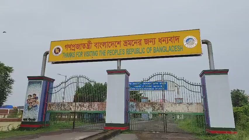 The video shows a close-up shot of a large yellow sign mounted above an entrance gate. The sign is written in both Bengali and English. The Bengali text reads "গণপ্রজাতন্ত্রী বাংলাদেশ ভ্রমনের জন্য ধন্যবাদ," and the English text reads "THANKS FOR VISITING THE PEOPLE'S REPUBLIC OF BANGLADESH." The sign has two circular emblems on either side, and a picture of a monument on the right side. Below the sign, there is a metal gate with decorative patterns. On the left side of the gate, there are colorful murals painted on a pillar, depicting a group of people. The overall scene suggests an entrance to a border crossing or a significant point of entry into Bangladesh. The sky in the background is overcast.