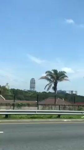 A car is traveling along a highway on a bright, sunny day. The sky is clear blue with a few white clouds. Along the roadside, there are palm trees, greenery, and a building. A tall, black, metal structure that appears to be a framework for a billboard is passed. A concrete wall lines the side of the road.