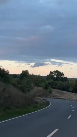 The video shows a winding road through a hilly, tree-lined landscape during twilight. The sky is overcast with patches of pale yellow and pink, indicating sunset or sunrise. A white dashed line marks the center of the asphalt road. A metal guardrail runs along the left side of the road. The vegetation is lush with various shades of green. There are no people or vehicles visible in the frame. The camera appears to be moving, giving the impression of travel.