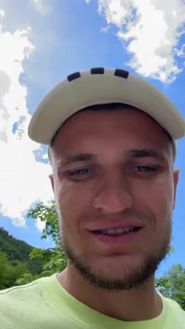 A close-up selfie-style video shows a young man with blue eyes and a light beard, wearing a white baseball cap with black stripes and a neon green t-shirt. He is outdoors with a blue sky and white clouds visible in the background, along with some green foliage. The man smiles slightly and appears to be speaking, though no audio is provided.