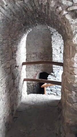 The video shows a narrow, stone passageway with an arched ceiling. There are wooden beams supporting the ceiling in the middle of the passageway. The walls are made of rough stone and brick. At the end of the passageway, there is a dark opening, possibly a doorway or another passage. The lighting is dim, suggesting an indoor or underground location. There are no people or significant objects visible in the scene.